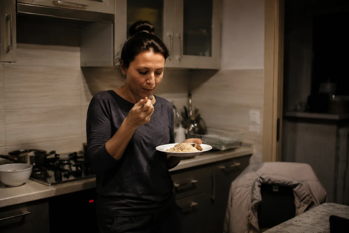 A woman eating standing at the kitchen counter late in the evening, a plate in her hand.