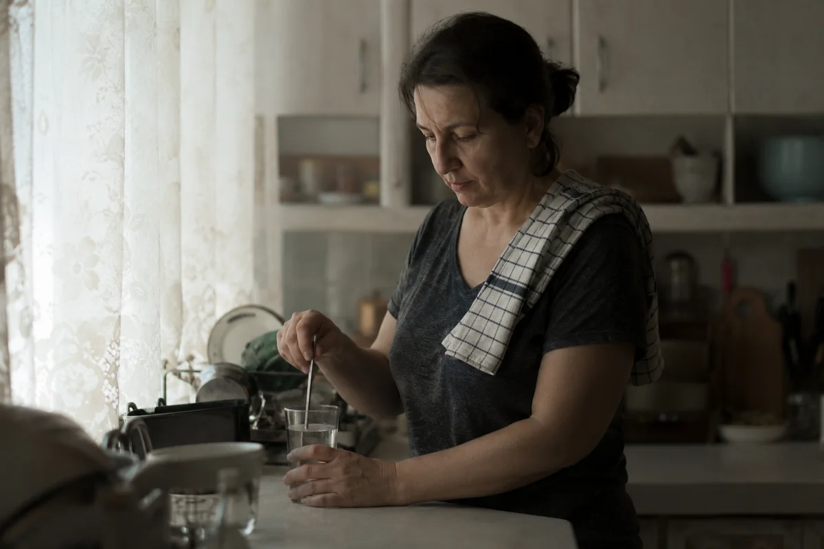 Жена бърка чаша вода с дълга лъжица до кухненския плот, сутрешна светлина отляво.